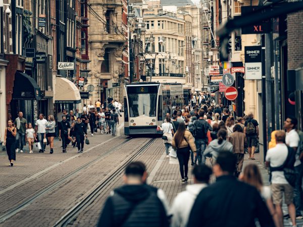 Bustling street scene in Amsterdam featuring a tram, shops, and diverse pedestrians on a lively day. Photo source: Metehan Demirkaya / Pexels.