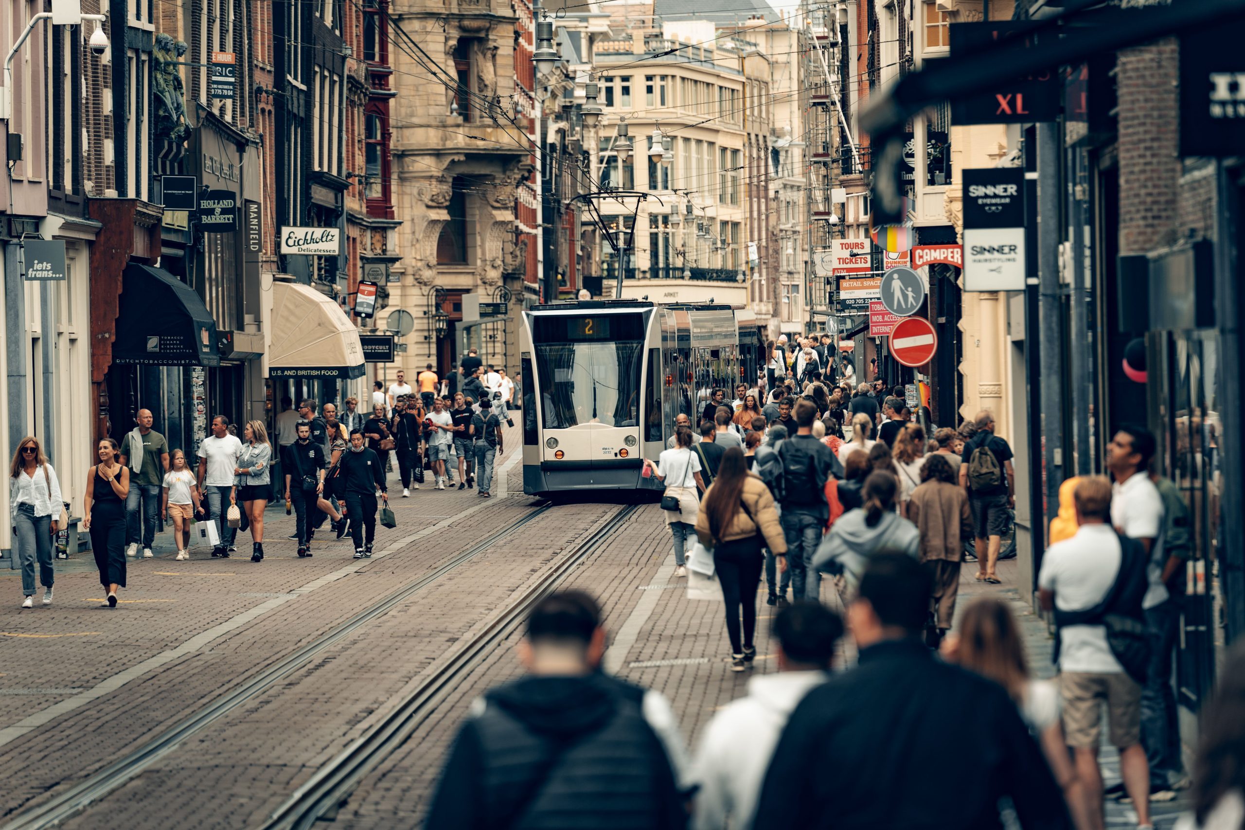 Bustling street scene in Amsterdam featuring a tram, shops, and diverse pedestrians on a lively day. Photo source: Metehan Demirkaya / Pexels.
